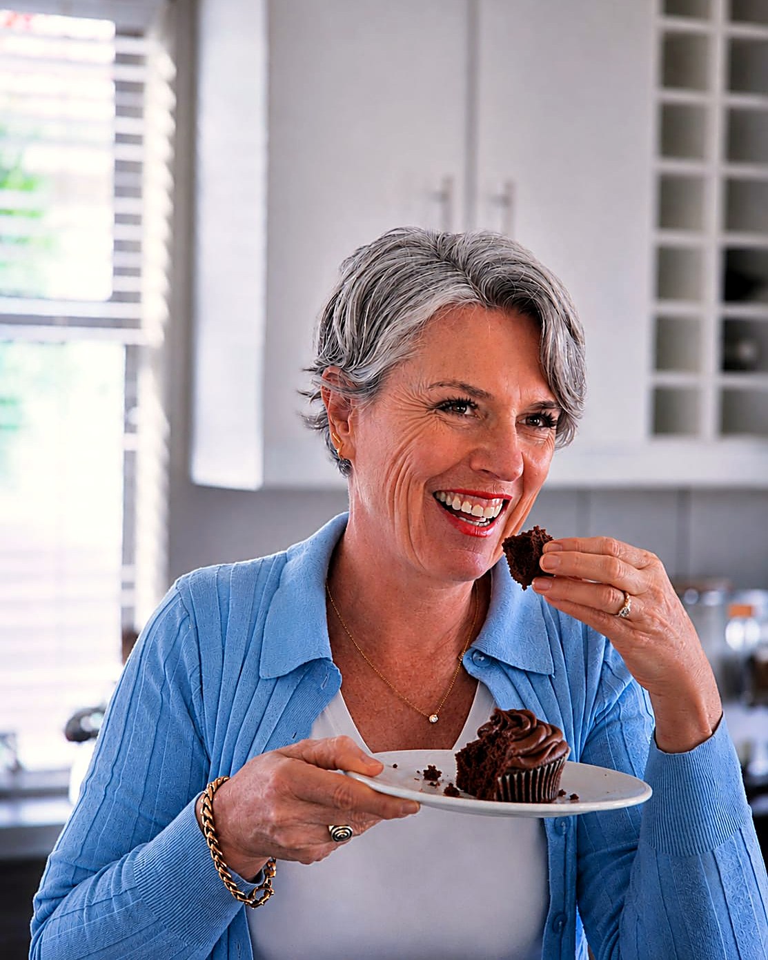 Sorea in her kitchen, smiling as she enjoys a chocolate cupcake she holds on a plate