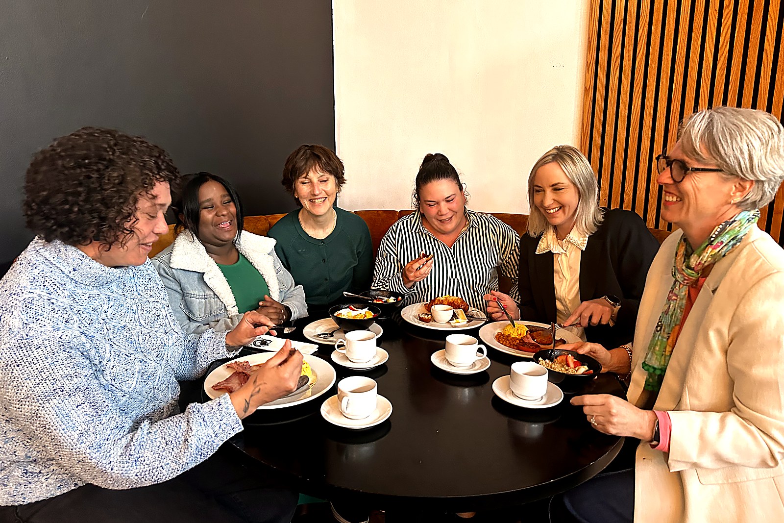 Sorea smiling with a diverse group of women around a caf&eacute; table, sharing breakfast and coffee together